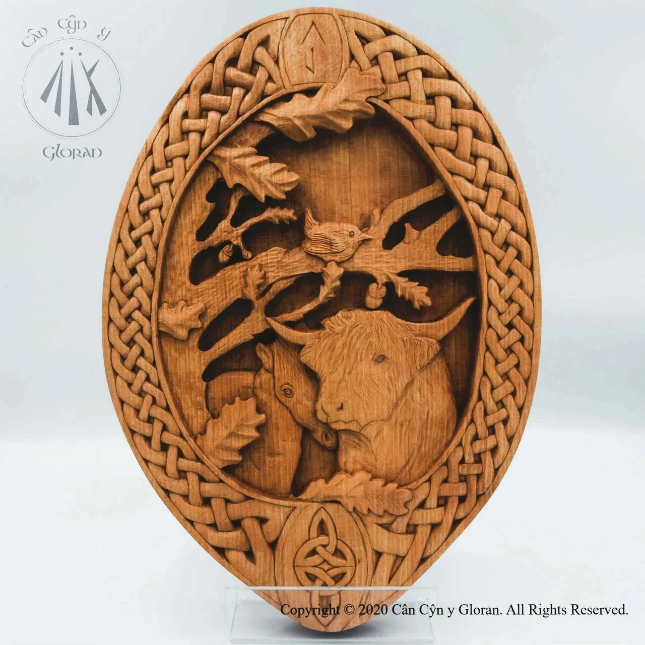 A work of art in beech wood. A wren sits in the centre, perched on the branch of an oak tree, while underneath a horse nuzzles into a highland cow. The scene is bordered by intricate Celtic knotwork, the entire thing carved by hand by a Welsh Gloran carver.