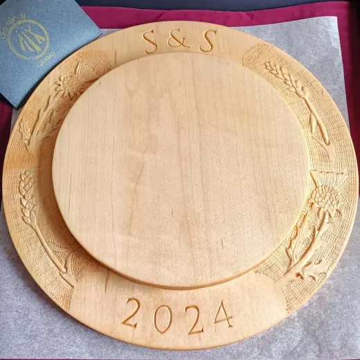 A maple breadboard with thistles, wheat sheafs, initials and the year 2024 carved into it